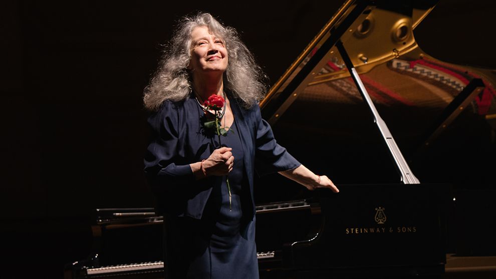 Frau mit langen grauen Haaren (Martha Argerich) steht mit Rose in der Hand vor einem Konzertflügel