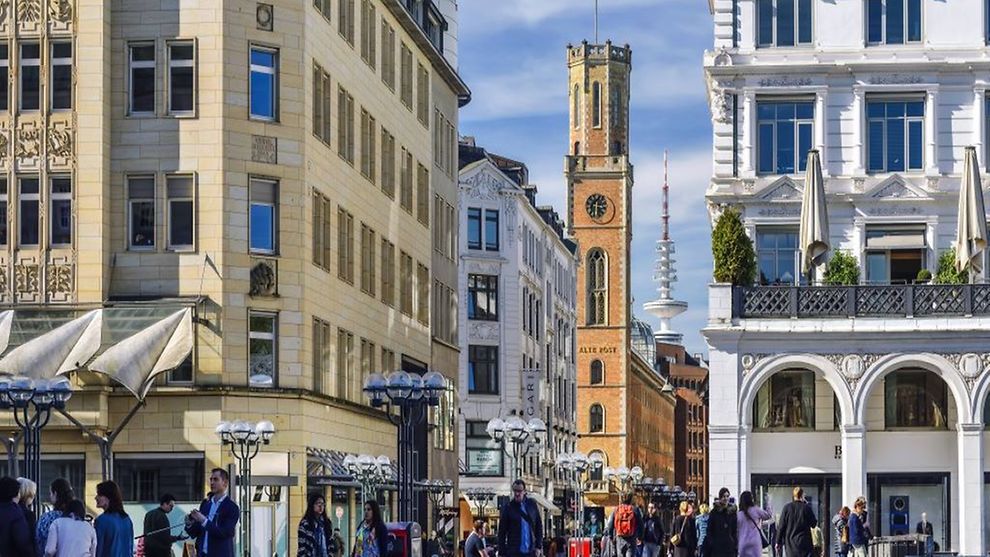  Auf dem Bild gehen Menschen in die Straße Große Bleichen. Im Hintergrund sieht man den Fernsehturm Hamburg, davor die Alte Post, die einen hohen Turm auf orangem Backstein hat.