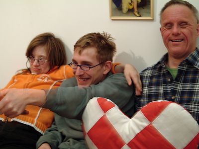  Drei Menschen mit Behinderung sitzen auf dem Sofa, einer hält eine Fernbedienung in der Hand.