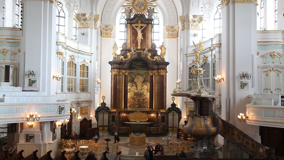 Im Innenraum der Kirche St. St. Michaelis sieht man einen großen Altar, weiße Wände und braune Holzbänke. Alles ist mit Gold und Malereien verziert. 