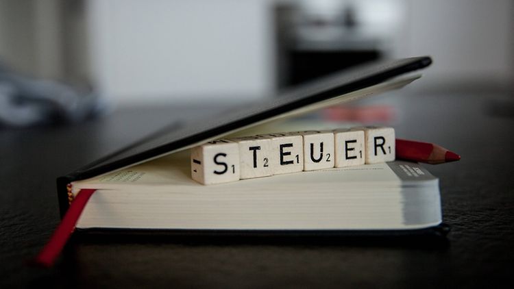  Ein ledergebundener schwarzer Kalender mit rotem Lesezeichenband liegt auf einem Tisch. Unter dem Deckel klemmen Scrabble-Würfel, auf denen das Wort "Steuer" zu lesen ist.