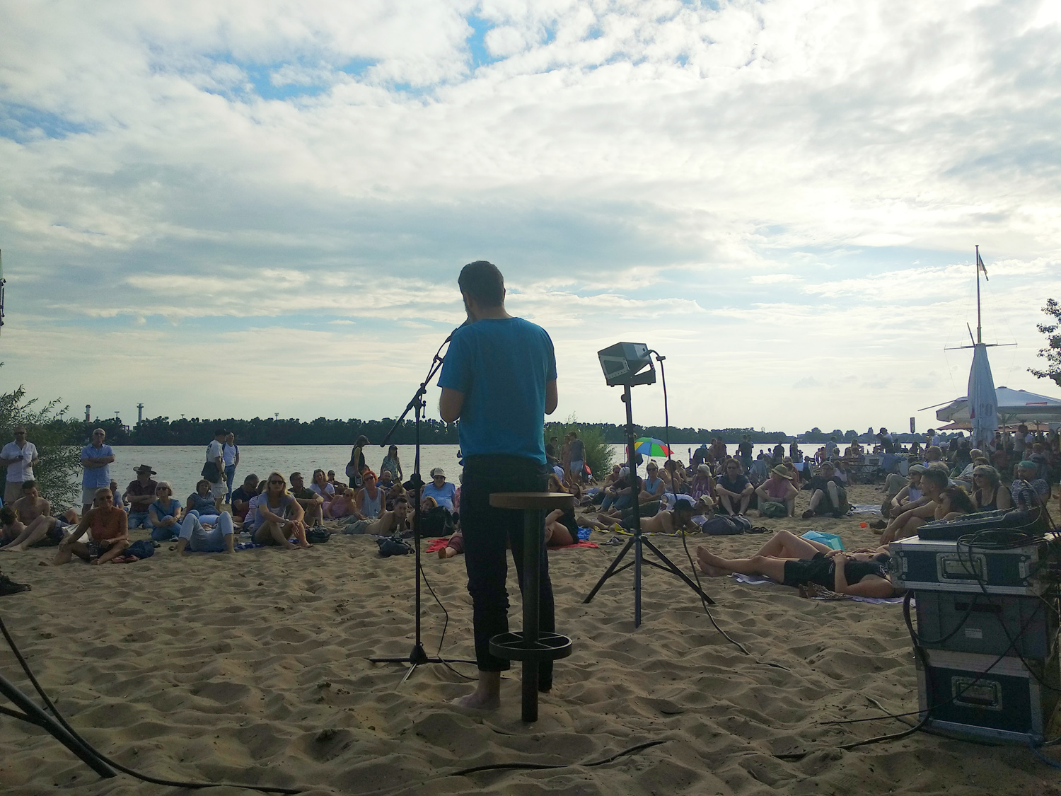 Ein Dichter im blauen T-Shirt liest am Strand an der Elbe vor Publikum, das im Sand sitzt