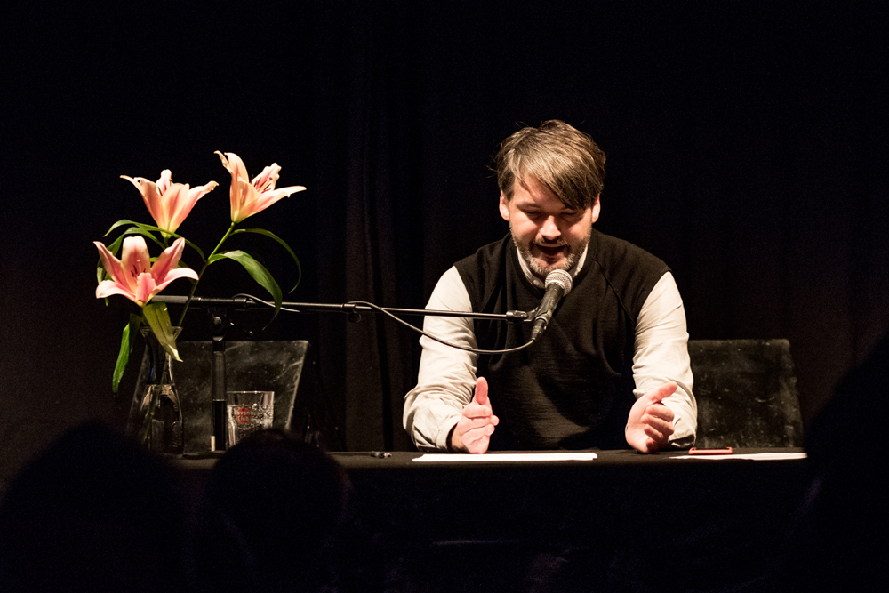 Mann sitzt an einem Tisch, vor sich ein Strauß Lilien, und liest aus einem Manuskript ins Mikrophon