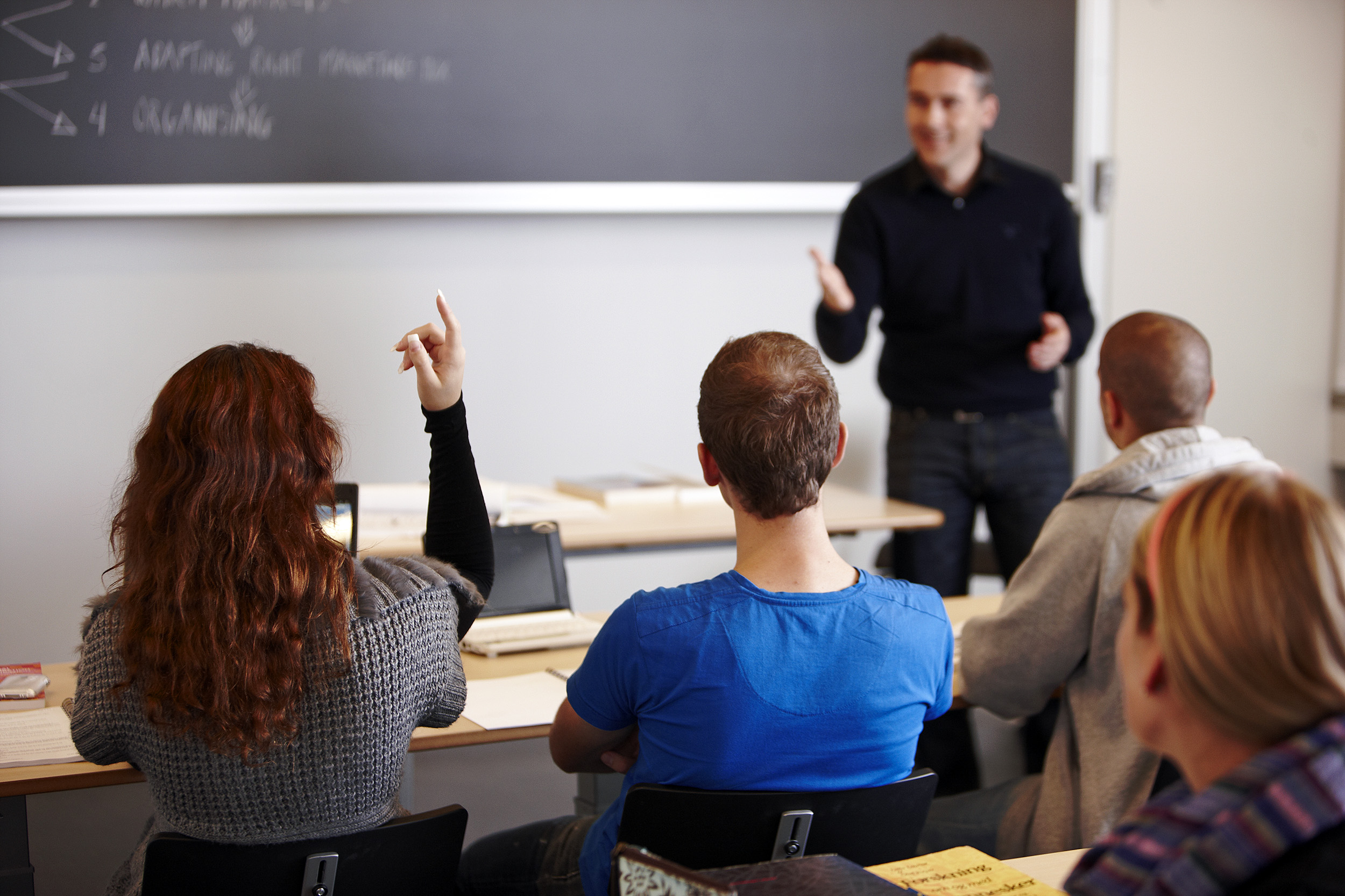 Ein Lehrer steht vor einer Tafel und nimmt eine Schülerin dran, die ihren Finger hebt. Drei weitere Schüler sitzen neben ihr.