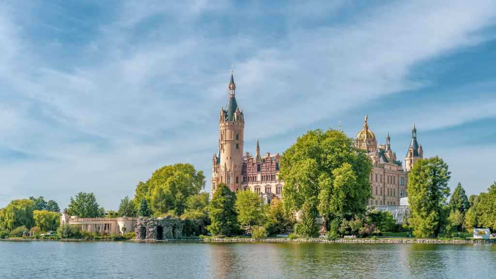 Das Schweriner Schloss steht auf einer Schlossinsel in einem See. Man sieht Bäume und die Spitzen des Schlosses.