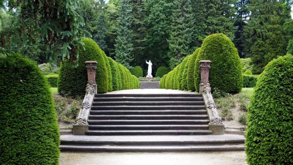 Eine breite Treppe führt auf einen Weg. Am Ende des Weges steht eine weiße Statue. Der Weg ist von Bäumen gesäumt.