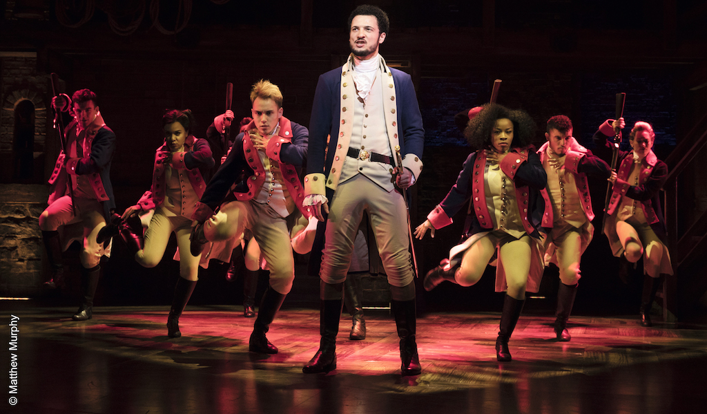 Szenenbild aus Hamilton: Alexander Hamilton uvd andere Castmember stehen in Uniform Spalier