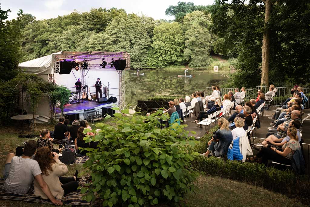 Eine Bühne mit zwei Künstlern umgeben von Bäumen und Wasser. Vor der Bühne sind viele Sitzplätze mit Publikum.