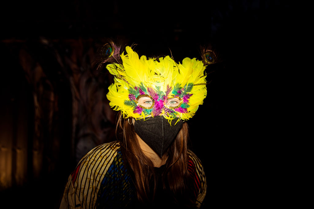 Eine Frau mit einer Vogelmaske aus gelben Federn und einer FFP2 Maske im Gesicht.