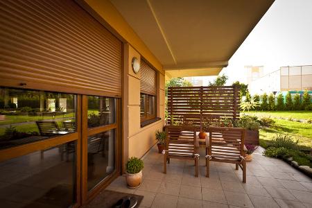 Eine Terrasse aus Holz mit Holzgestühl und Außenrollos an der Fensterfront