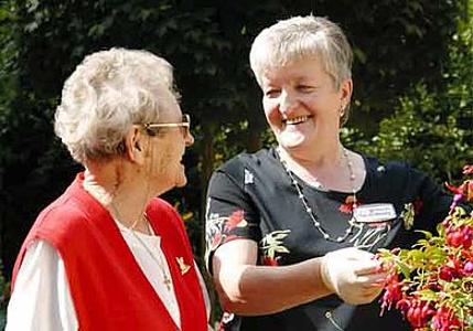Zwei ältere Frauen stehen vor einem Blumenbusch und unterhalten sich