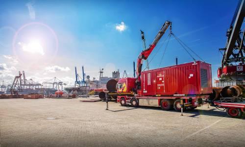 Roter LKW mit Kran auf einem Hafengelände, im Hintergrund Container und Hafenkräne