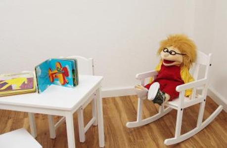 Weißer Kindertisch mit Stuhl davor und Kinderbücher auf dem Tisch, daneben kleiner Schaukelstuhl mit Puppe darauf.