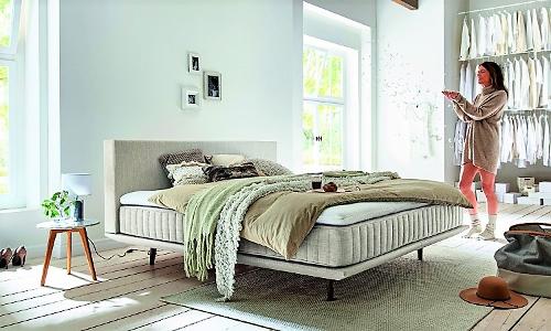Ein Bett steht auf einem hellen Teppich an einer hellen Wand zwischen zwei Fenstern