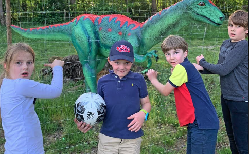 Vier Kinder mit einem Fußball stehen vor einer Dinosaurierfigur