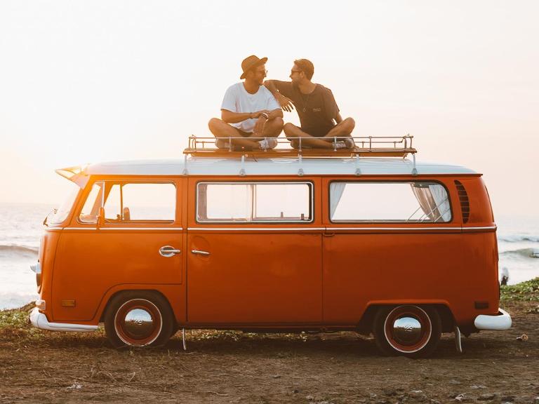 Seitenansicht auf einen orangefarbenen VW Bully Reisebus, mit zwei Männern auf dem Dach sitzend, vor einem Sonnenuntergang