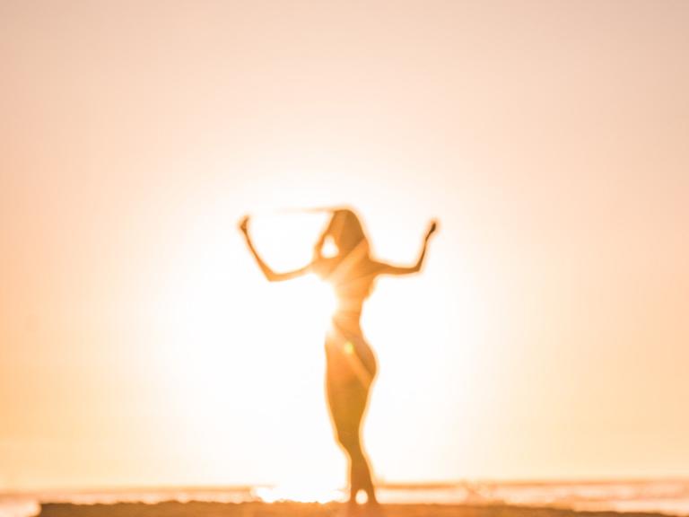 Frau geht am Strand, hebt die Arme in die Luft und wird von der Sonne angestrahlt