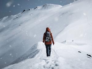 Person mit roter Winterjacke und einem Rucksack auf dem Rücken geht durch den Schnee auf einen Berg zu 