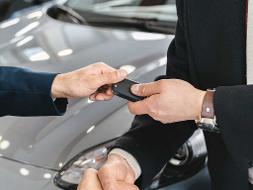 Eine Männerhand übergibt einer anderen Männerhand einen Autoschlüssel, im Hintergrund ein schwarzes Auto