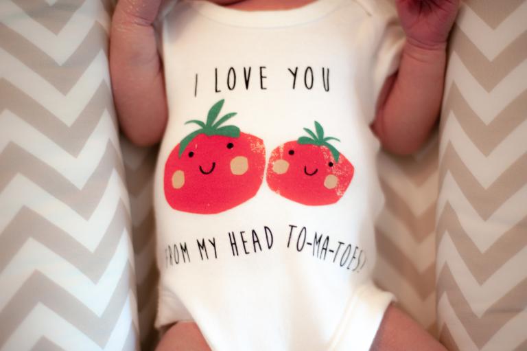 Ein Baby liegt auf einer gemusterten Decke und hat einen weißen Strampler mit zwei roten Tomaten darauf an auf dem steht I LOVE YOU FROM MY HEAD TO-MA-TOES!