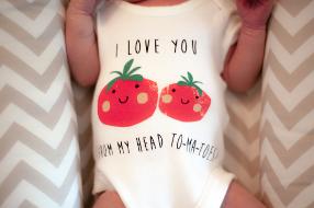 Ein Baby liegt auf einer gemusterten Decke und hat einen weißen Strampler mit zwei roten Tomaten darauf an auf dem steht I LOVE YOU FROM MY HEAD TO-MA-TOES!