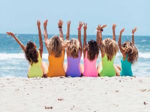 Vier Frauen sitzen mit dem Rücken zur Kamera am Strand, blicken auf das Wasser und heben die Arme in die Luft- jede hat einen andersfarbigen Badeanzug an