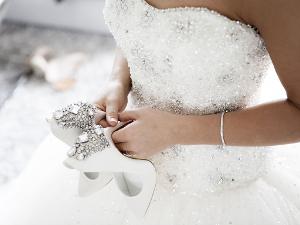 Eine Frau hat ein schulterfreies Hochzeitskleid mit Glitzersteinen besetzt an, weiße Brautschuhe in der Hand und ein silbernes Armband um