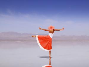 Eine Frau mit einem orangefarbenen Hut auf dem Kopf, einem weißen Spaghettiträgetop und einem orangefarben Rock steht auf einem Bein am Strand, das linke Bein nach rechts hebend und die Arme in der Luft