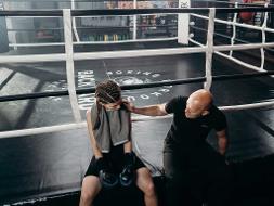 Eine Frau und ein Mann sitzen am Rand eines Boxrings, er legt den Arm auf ihre Schulter und spricht zu ihr