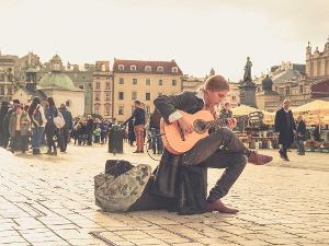 Ein Straßenmusiker sitzt auf einer Box auf einem öffentlichen Platz, im Hintergrund sieht man Menschen und Häuser