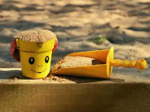 Ein gelber Eimer mit einem aufgemalten Gesicht und eine gelbe Schaufel liegen auf dem Rand einer Sandkiste