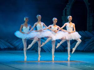 Vier Personen in weißer Ballettkleidung stehen nebeneinander, jeder auf einer Fußspitze und halten sich die Hände