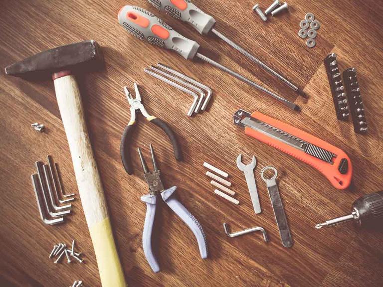 Hammer, Cuttermesser, Schrauben, Dübel, Zangen und Schraubenzieher liegen auf einem Holzuntergrund