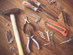 Hammer, Cuttermesser, Schrauben, Dübel, Zangen und Schraubenzieher liegen auf einem Holzuntergrund