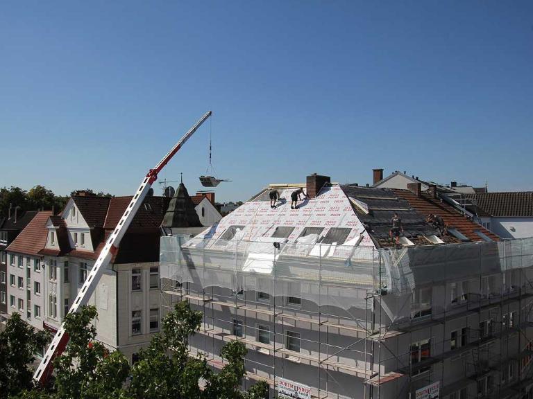Menschen arbeiten auf dem Dach eines Hauses, ein Kran bringt das Material nach oben