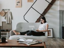 Eine Frau sitzt auf einem Sofa die einen Laptop auf ihren Knien stehen hat und daran arbeitet