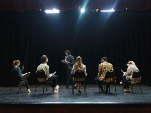 Fünf Menschen sitzen auf einer Bühne auf einem Stuhl- ein Mensch steht vor ihnen und hält ein Stück Papier in der Hand