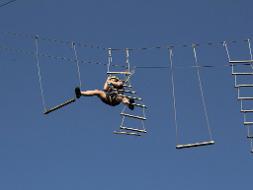 Ein Mann klettert in der Luft, vor blauem Himmel an Seilen mit Holzstücken herum