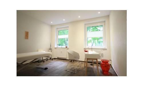 Behandlungszimmer mit weißen Wänden, Liege, Schreibtisch mit zwei Stühlen und zwei Fenstern