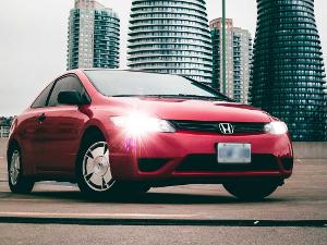 Ein roter Honda hat die Scheinwerfer an und steht schräg vor der Kamera, im Hintergrund Hochhäuser