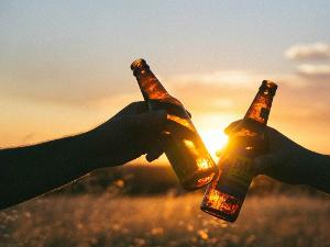 Zwei Hände halten jeweils eine Bierflasche in der Hand und stoßen an- im Hintergrund ein Sonnenuntergang
