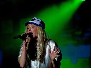 Eine Frau mit langen, blonden Haaren und einem Cap auf dem Kopf- singt in ein Mikrofon, im Hintergrund ist grünes Licht und Nebel