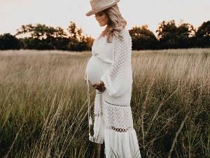 Eine schwangere Frau, mit weißer Kleidung und einem weißen Hut auf dem Kopf steht in einem Feld aus Gräsern