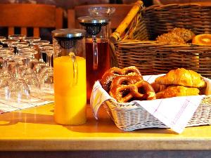Gläser stehen umgedreht auf einem Holztisch, daneben Glasbehälter mit Orangensaft und Tee, sowie ein weißer Brotkorb mit Laugenbretzeln und Croissants darin
