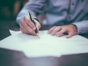 Ein Mann in einem blauen Hemd sitzt an einem Tisch auf dem Unterlagen liegen die er mit dem Stift in der Hand unterzeichnet