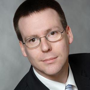 Portraitfoto eines Mannes mit Kurzhaarschnitt, einer Brille, einem weißen Hemd, Krawatte und einem dunklen Jackett 