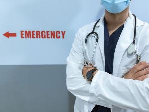 Arzt mit weißem Kittel und verschränkten Armen steht vor einer Wand mit der Aufschrift Emergency