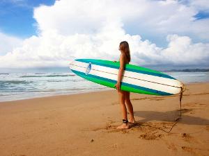 Ein Mensch hält das Surfbrett unter dem Arm und steht am Strand, mit Blick auf das Meer