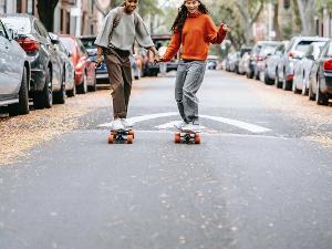 Zwei Menschen auf jeweils einem Skateboard fahren nebeneinander auf einer Straße und halten sich an den Händen