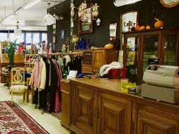 Eine Holzkommode, Spiegel, ein Teppich und eine Kleiderstange mit Kleidung auf den Bügeln hängend stehen in einem Geschäft